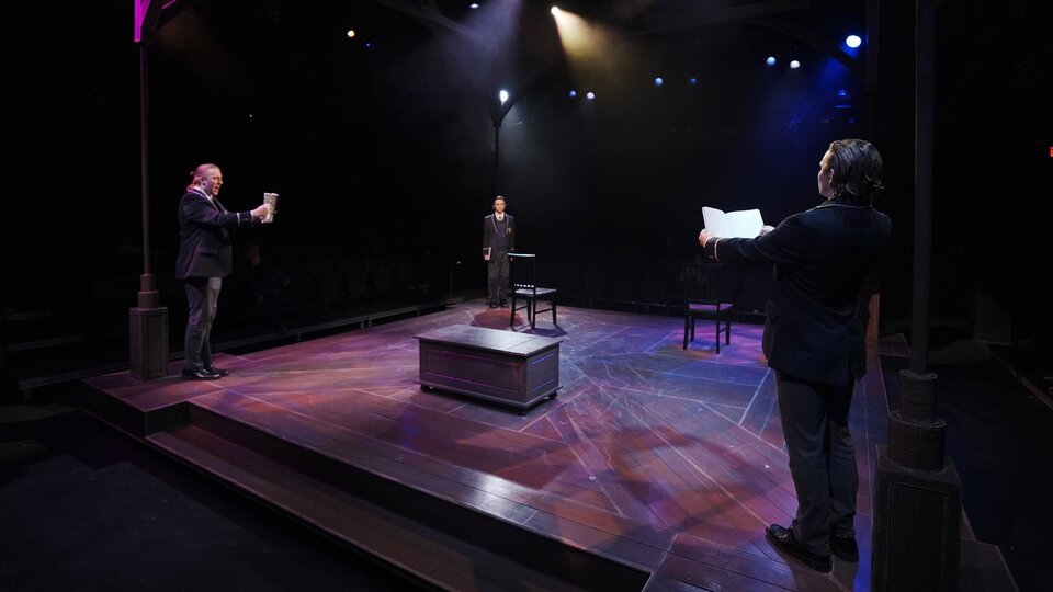 Three actors stand at the edge of a stage in a triangular formation reading lines.
