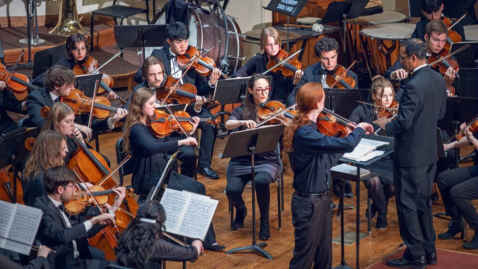 A wide shot of an orchestra playing with a long red-haired violin soloist standing and playing.