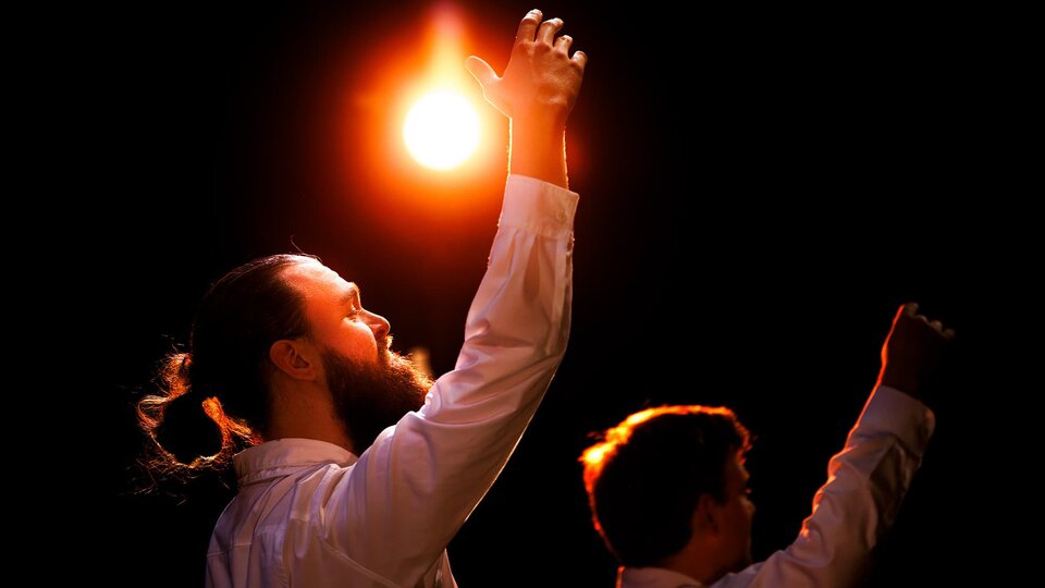 Two actors on a darkened stage reach their rights arms up, a single light casts an orange glow.