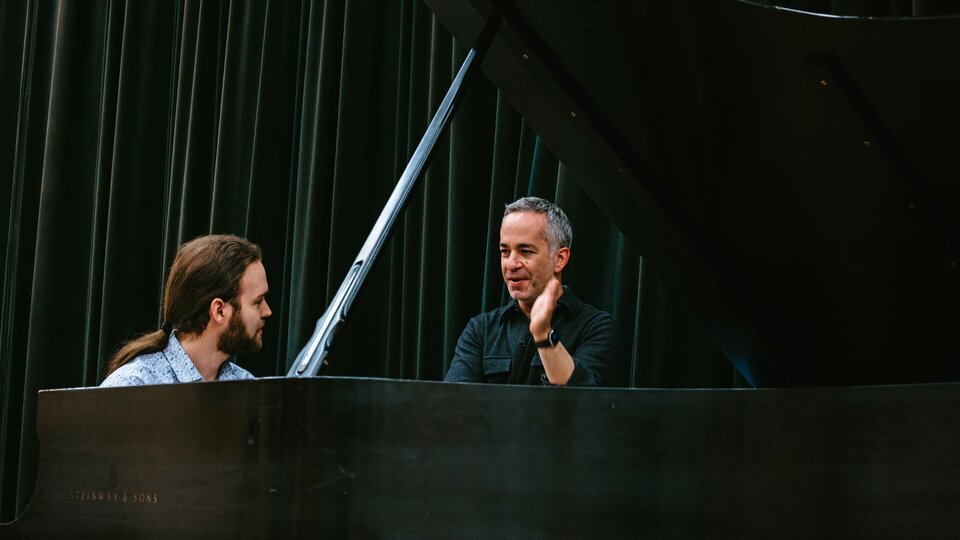 A student sitting at a piano looks at a guest artist giving them feedback.