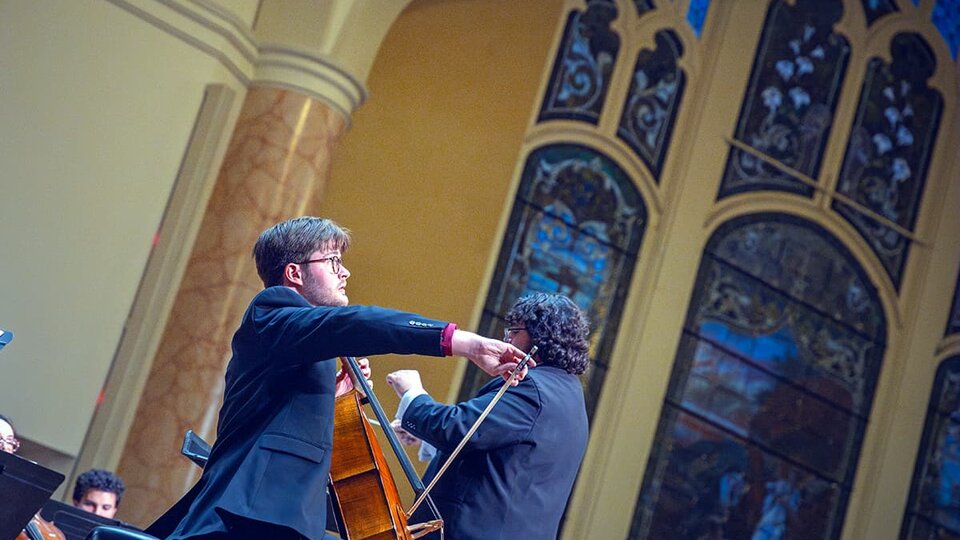 A celloist plays on stage with huge stained glass windows in the background.