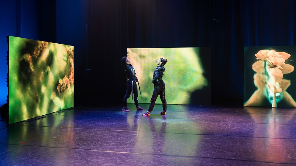 Two dancers in motion capture suits performing on stage.