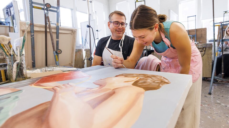 A Student works on a painting while an instructor looks on.