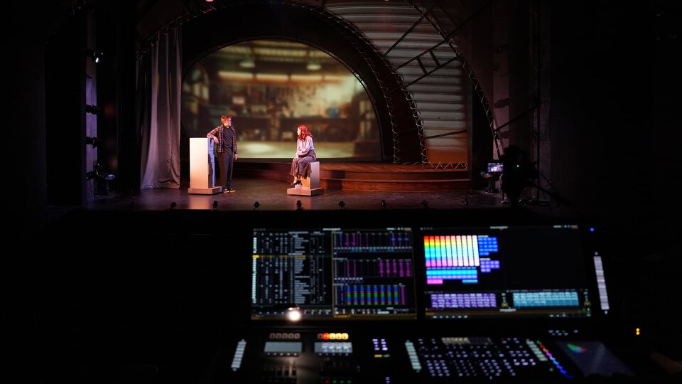 Two actors block out a scene on stage while the theatre control panel sit alight within the darkened theatre.