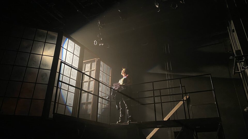 A crew member stands in the particles of the beam of the spotlight as they stand on a catwalk.