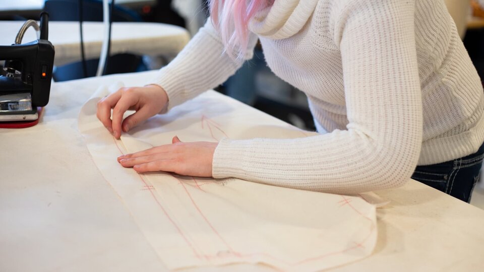 A person in a white sweater leans over a work table arranging a sewing pattern.