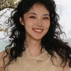 Smiling woman with curly black hair outdoors near a tree, wearing a beige top.