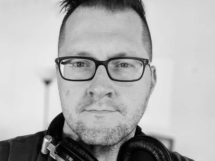 Man wearing glasses with headphones around his neck, in black and white.