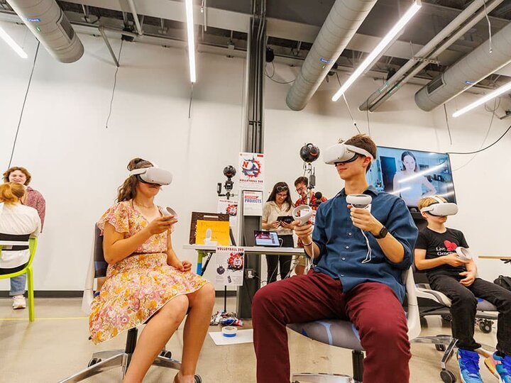A few people sit in chairs wearing virtual reality googles with hand controllers.