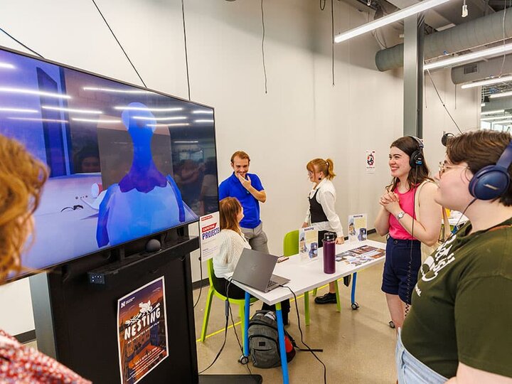 A few people stand with headphones on watching a large screen TV.