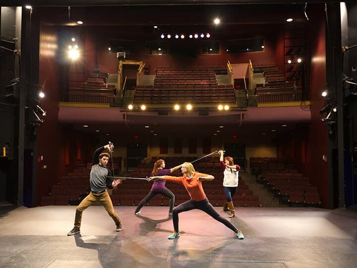 Actors practice stage combat with empty theatre seats in the background.