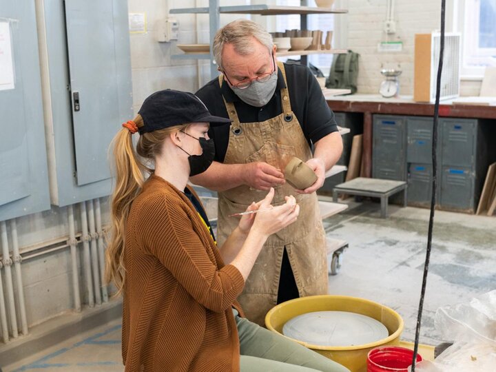 Professor Pete Pinnell holding and looking over a ceramic work a School of Art student who is sitting at a wheel in the ceramics studio is working on.
