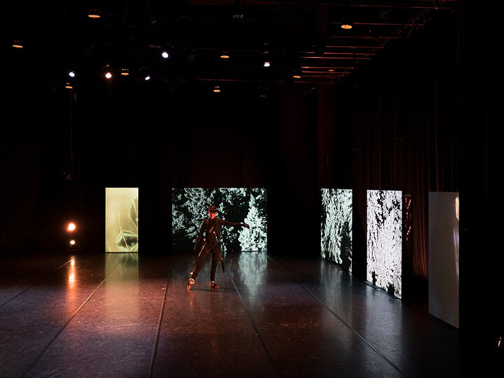 A dancer stand in pose in the center of a dark stage with projected panels surrounding it.