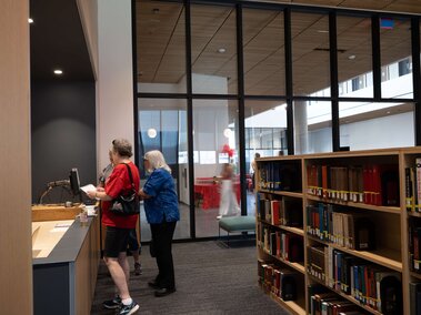 Library interior with people at a counter, bookshelves, and glass walls leading to another area.
