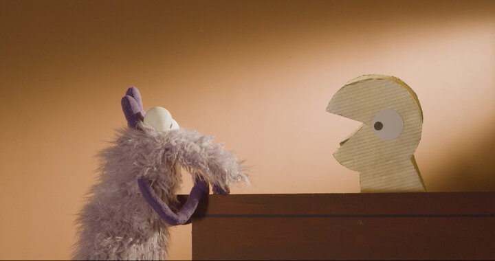 Purple furry puppet "Delmar" faces cardboard character on a wooden surface, warm orange background.