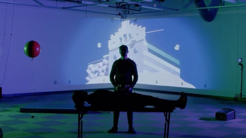 A silhouetted figure standing in front of a table with another laying upon it with a projected background on the wall behind them. 