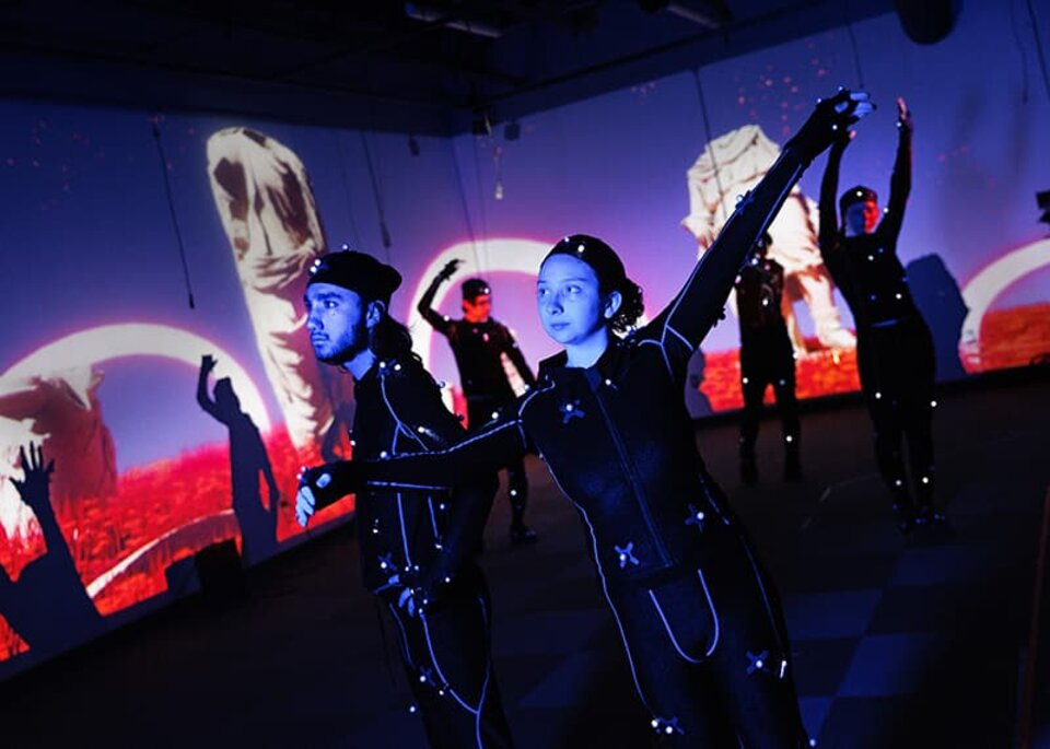 A group of students in motion capture suits perform on a darkened stage with projected backgrounds on the walls surrounding them.