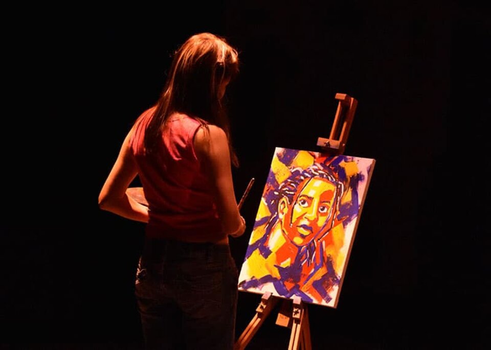 A woman stands on a dark stage working on a painting on an easel. 