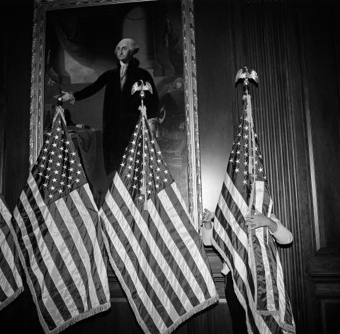 An aide to House Speaker Nancy Pelosi adjusts a row of flags in front of a painting of George Washington at the U.S. Capitol, preparing for Democratic leaders to speak after the vote on December 18, 2019, to impeach President Donald Trump the first time. The House impeached the president for abuse of power and obstruction of Congress for trying to get Ukraine to investigate Joe Biden. © Louie Palu. 