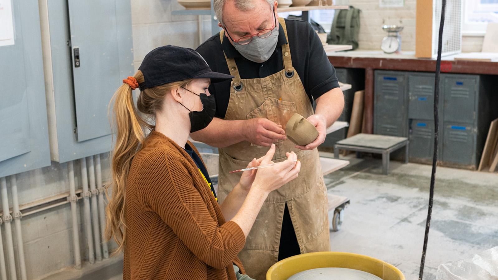 Professor Pete Pinnell holding and looking over a ceramic work a School of Art student who is sitting at a wheel in the ceramics studio is working on.