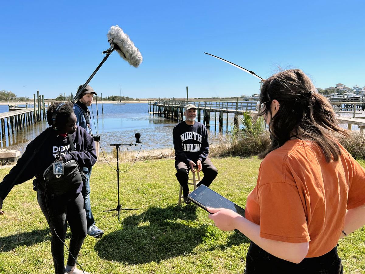 Three Emerging Media Arts students on an outdoor film shoot on-site with a resident of Seabreeze sitting on a stool in front of a shore with docks.