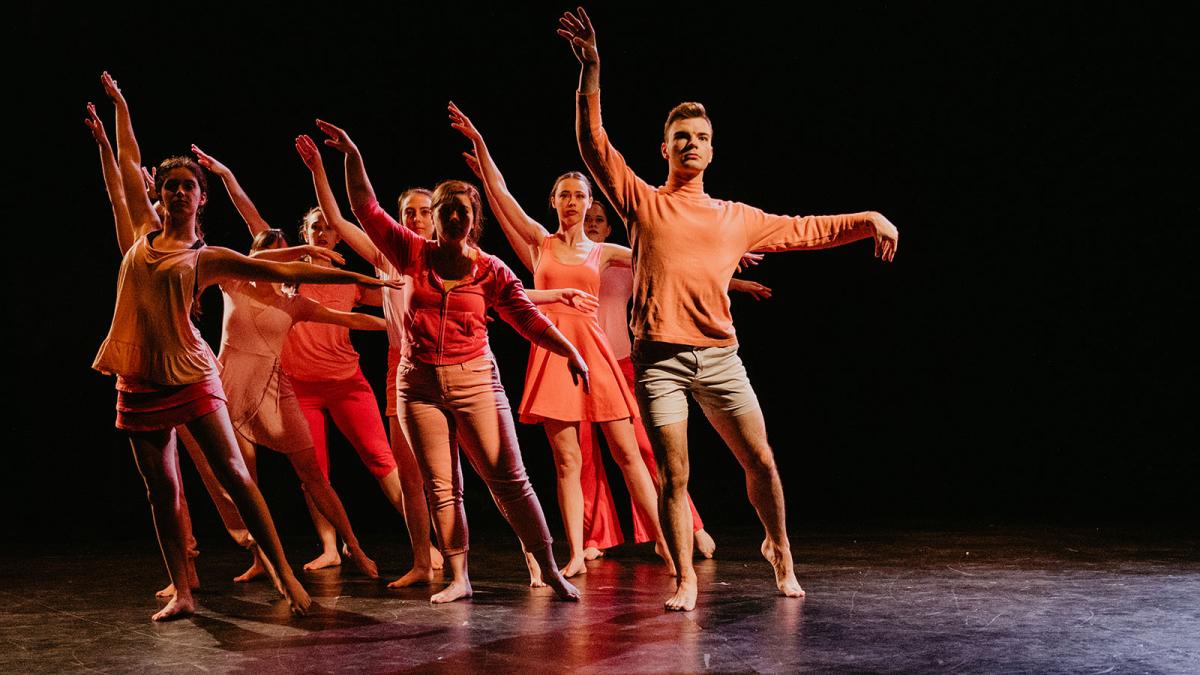 A group of dancers on stage all wearing outfits in various shades of orange in a  pose synced with the dancer at the the front of the group.