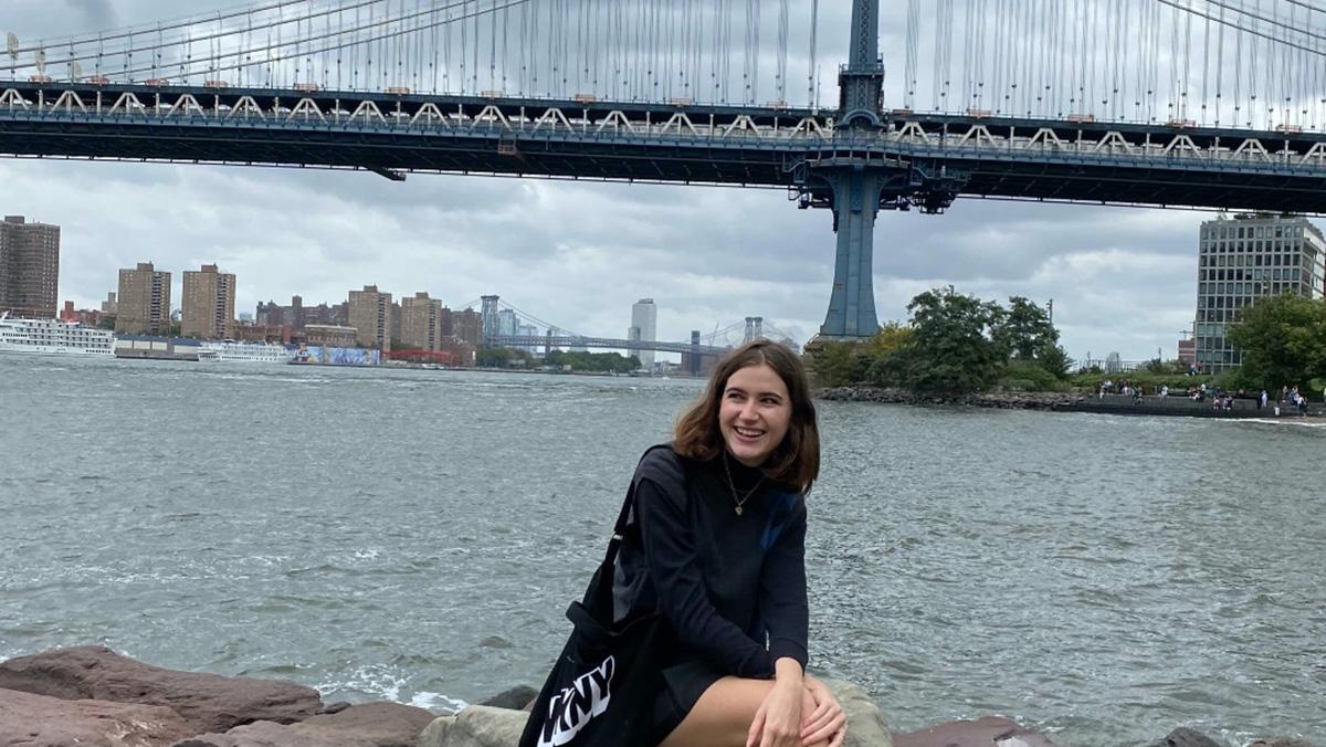 Sabrina Sommer, smiling and looking off-frame while sitting on a large rock in front of a body of water and bridge in New York City.  