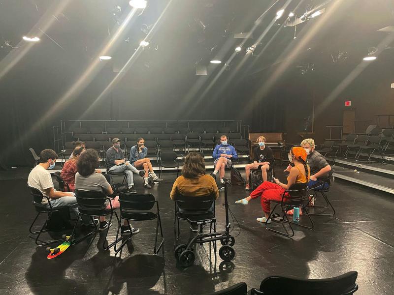 A circle of Carson School students in a black box theatre room facing each other and engaging in conversation.