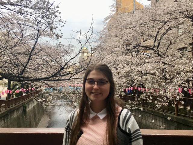 Mikayla Zulkoski in Japan on a bridge with a waterway lined with cherry trees in full bloom behind her.