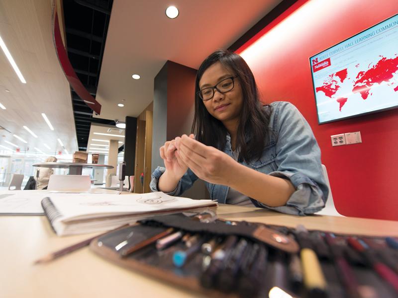 Graphic Design alumna Emily Tran sharpening a pencil while sitting at a table in an open study space with drawing supplies and drawing pad in front of her.