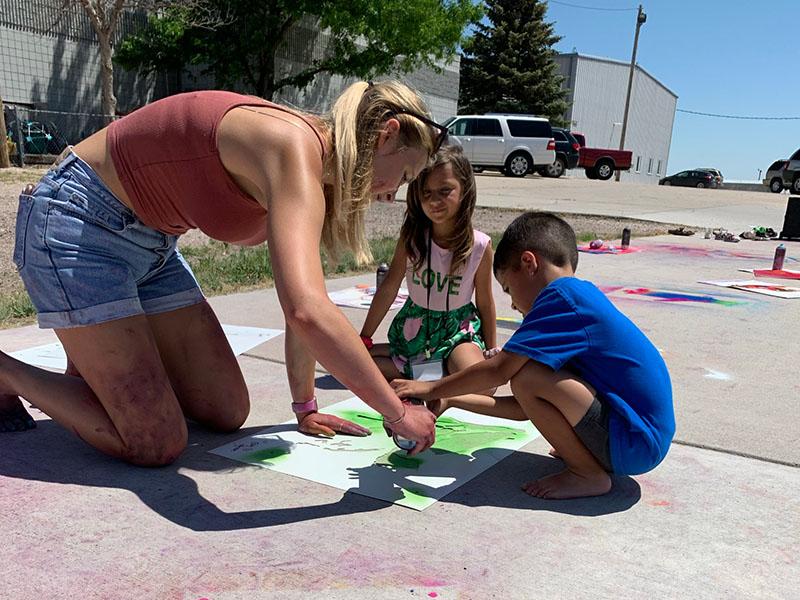 School of Art student outdoors kneeling next to two young people creating sidewalk art in a rural community.