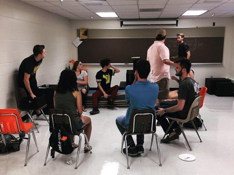 A group of Nebraska students in the Lazzi Improve Troupe sitting in a half-circle while a few of their peers play out an improv scene.
