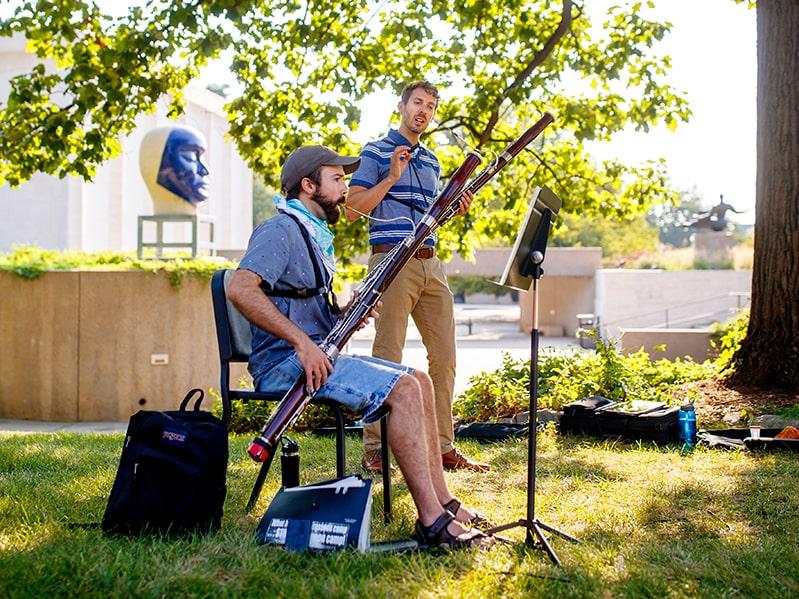 Glenn Korff School professor and student outdoors on a sunny day under a shady tree in the sculpture garden. The student is sitting in a chair, playing a bassoon in front of a music stand while the professor gives instruction.