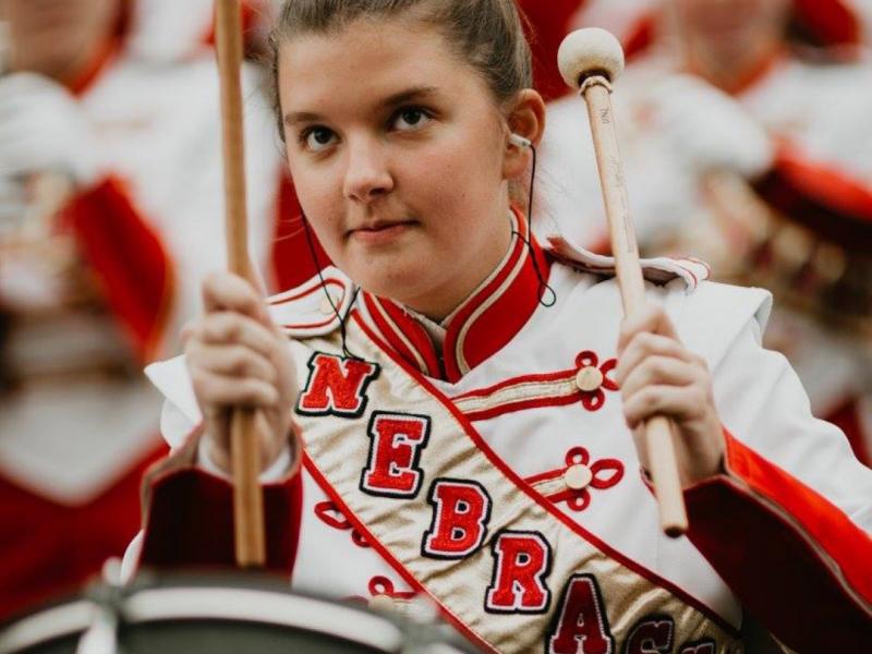 Music Education alum Alyssa Soppe in marching band regalia playing the drum with large drumsticks in hands.