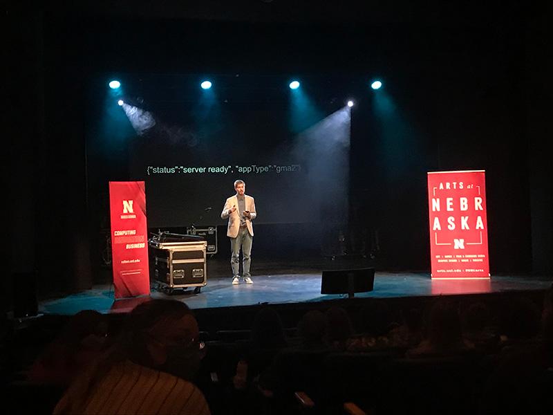 Theatre student giving a presentation on a lit stage with Nebraska banners on either side of him and a projected slideshow behind him.